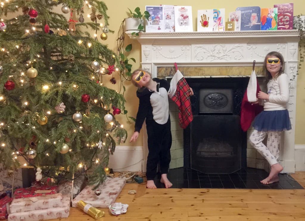 Children beside a decorated Christmas tree and a fireplace hung with stockings